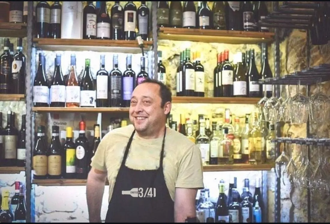 Tartufo, l'oste, sorridente davanti agli scaffali di bottiglie